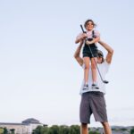 A smiling dad carrying his daughter on his shoulders as she holds a golf club outdoors