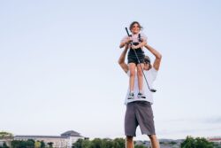 A smiling dad carrying his daughter on his shoulders as she holds a golf club outdoors
