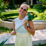 A smiling young woman carrying mini golf putters over her shoulder on a sunny outdoor course